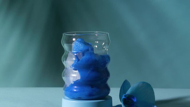 Vivid blue spirulina matcha milk pouring from pitcher over crystal ice into wavy bubble glass beside orchid on teal surface in slow motion