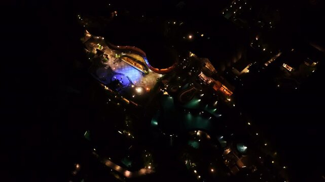 A dramatic high-altitude night drone shot reveals a resort's winding illuminated pools glowing against the surrounding darkness, with scattered garden lights dotting the grounds. Bali, Indonesia