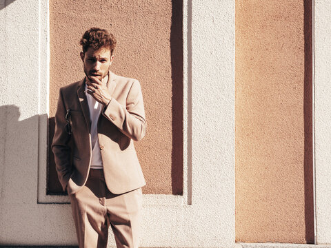 Handsome confident stylish hipster lambersexual model.Sexy modern man dressed in elegant beige suit. Fashion male posing in the street background in Europe city at sunset. In sunglasses