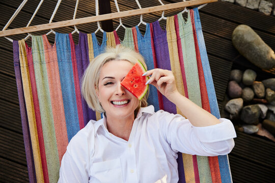 Playful mature woman holding watermelon slice over eye in hammock