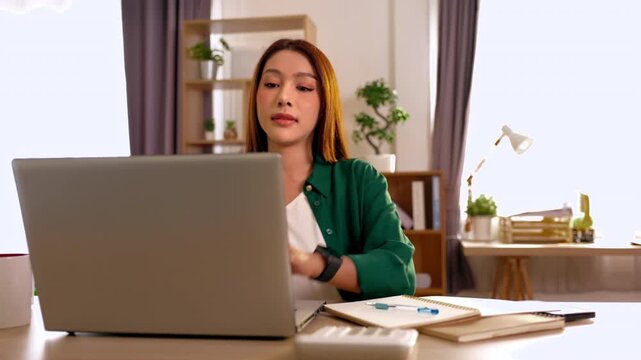 Young adult woman working on laptop at home office desk in modern remote work workspace, writing notes in notebook with pen, focused indoor daylight online learning study setup for office productivity