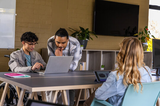 Diverse coworkers pointing and leaning at silver laptop on long wooden table, wheelchair present