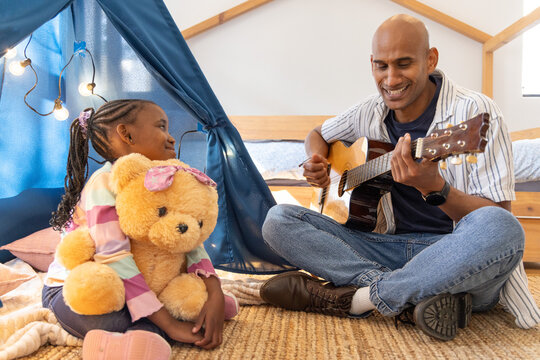 Asian father, African American child playing guitar at home holding teddy by play tent, copy space