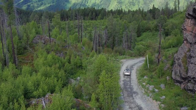 Aerial view of car driving along winding mountain road through forested valley with rocky cliffs in Katon Karagay national park, East Kazakhstan. Remote travel, wild nature and scenic landscape.