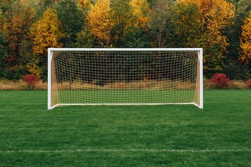 Fototapeta premium White soccer goal net stands on vibrant green sports field with backdrop of colorful autumn trees in park setting. High quality photo