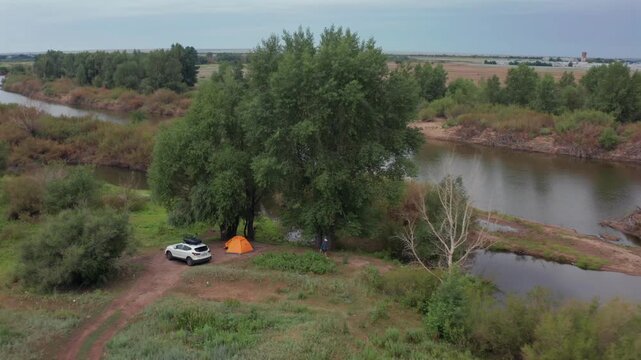 Aerial drone view of campsite with SUV and orange tent near Ilek River in Aktobe region Kazakhstan, green riverside trees, rural landscape, peaceful outdoor travel scene, nature and recreation