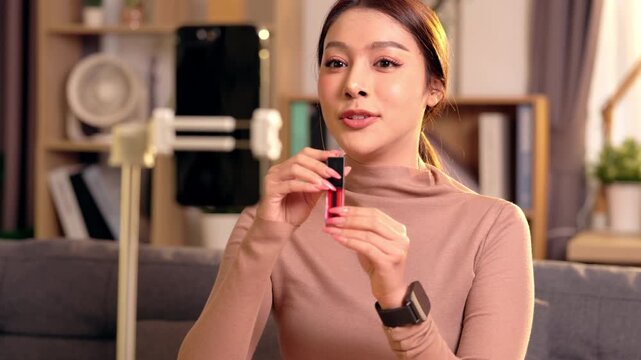 Woman using smartphone tripod for livestream makeup beauty tutorial home living room, young adult influencer smiling, waving video call, recording and presenting lipstick cosmetic tips warm light