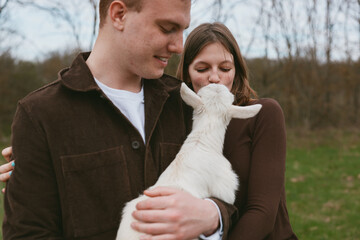 Fototapeta premium A couple outdoors with a gentle goat kid, sharing affectionate moment in a rustic countryside setting, showcasing lifestyle, tenderness, bonding, and calm, as the animal nuzzles close.