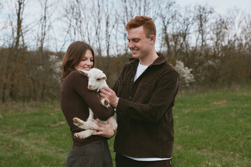 Fototapeta premium A happy couple outdoors with a small white goat kid, smiling as they cradle the playful animal in a green field, showcasing rural lifestyle and gentle interaction between humans and livestock.