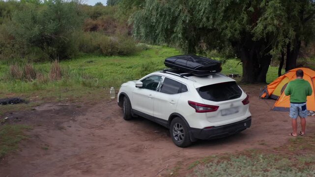 Traveler standing near orange tent and SUV with roof box at riverside campsite, green trees and natural surroundings, outdoor camping during road trip, peaceful holiday in Aktobe region Kazakhstan