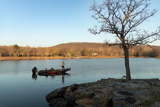 Lone man casting a fishing line from a boat near a rocky shoreline