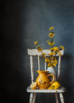 Still life with chair, jug, vase of yellow flowers against blue wall.