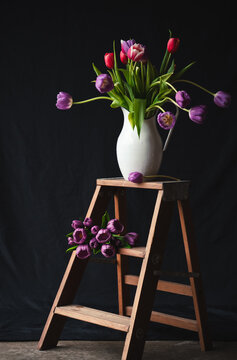 Jug of tulips on small wooden step ladder against black backdrop.