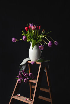 Jug of tulips on small wooden step ladder against black backdrop.