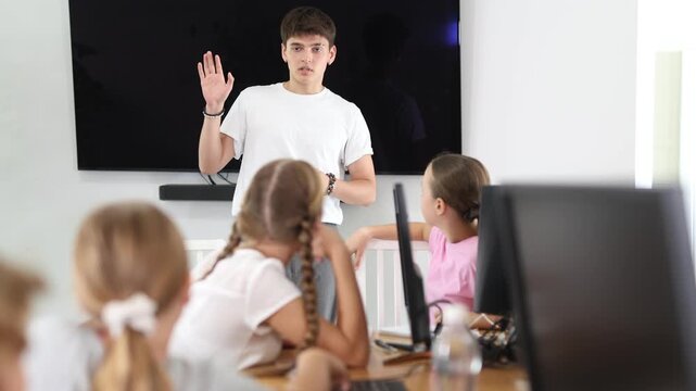 Young computer science teacher standing near an interactive whiteboard tells new material to teenage students in a school computer class