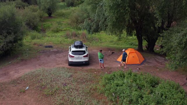 Drone view of travelers setting up orange tent beside SUV near Ilek River in Aktobe region Kazakhstan, green trees and dirt clearing, outdoor camping during road trip, peaceful natural environment