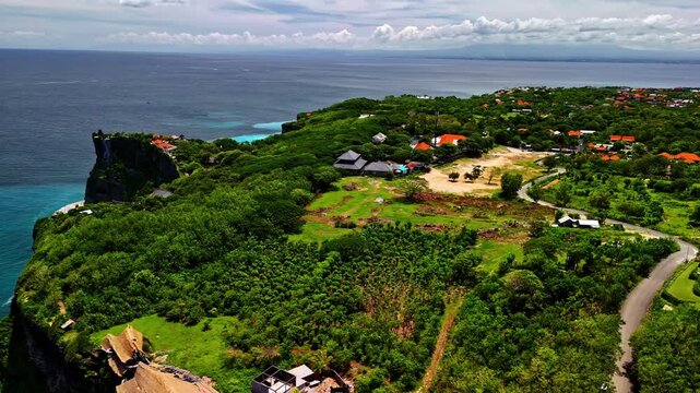 Panoramic drone aerial reveals the Uluwatu temple cliff complex perched above the Indian Ocean, surrounded by lush green vegetation, orange-roofed villas, and winding roads stretching to the horizon.