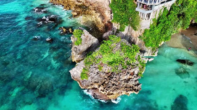 A striking aerial of a massive vegetation-crowned limestone sea stack rising from crashing turquoise waves, with a cliffside bar and terrace perched dramatically on the adjacent headland. Uluwatu,Bali