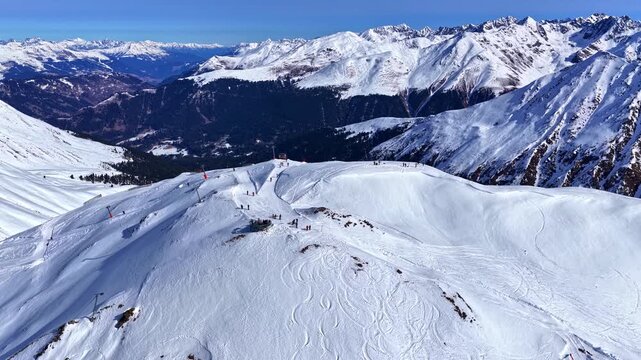 Sweeping aerial panorama over groomed ski slopes with skiers and dramatic snow-covered peaks under blue sky. Nauders, Austria