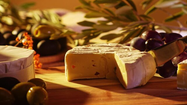 Close up of a hand slicing soft camembert cheese on a rustic wooden board surrounded by grapes olives and olive branches in warm golden light