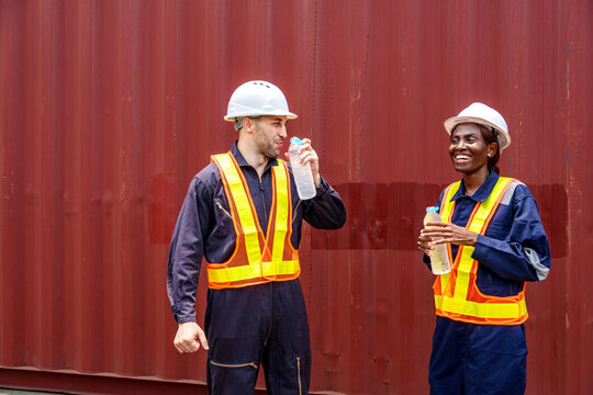 Smiling mid-adult Caucasian male and Black female industrial workers sharing a laugh while drinking water at an Asian container yard.