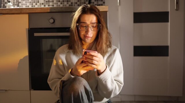 Young woman sitting on kitchen floor absorbed in smartphone addiction constantly checking screen unable to stop scrolling losing time and focus in warm evening home atmosphere