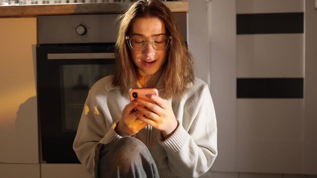 Young woman sitting on kitchen floor deeply addicted to smartphone unable to stop scrolling losing control over time and attention ignoring real life in warm evening home atmosphere