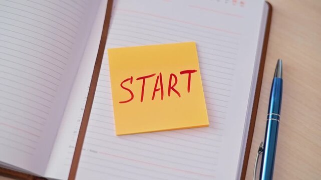 A hand places a sticky note that says start on an empty notebook on a desk. A pen is positioned nearby as a reminder to initiate tasks and ideas for future projects