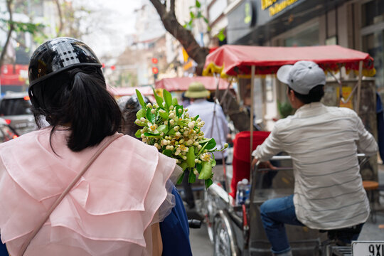 A woman wearing helmet carries fresh pomelo blossoms on a busy Hanoi street beside a passing cyclo vendor in soft daylight.