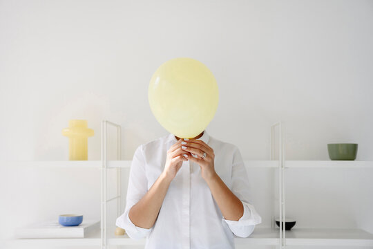 Woman holding a yellow balloon to hide face in bright white room