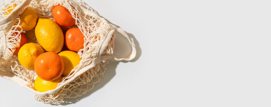 Lemons and tangerines in eco-friendly shopping bag on light gray background, flat lay top view. Mesh cotton string bag filled with citrus fruits. Concept of eco-friendly living, zero waste