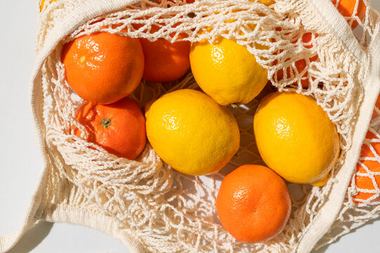 Lemons and tangerines in eco-friendly shopping bag on light gray background, flat lay top view. Mesh cotton string bag filled with citrus fruits. Concept of eco-friendly living, zero waste