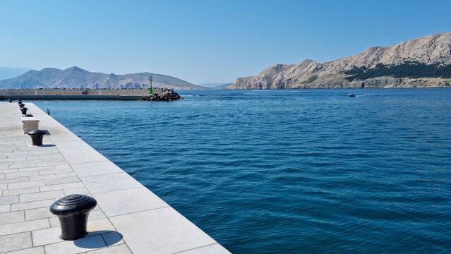 A paved stone pier with black mooring bollards leads the eye toward the deep blue Adriatic Sea and the rocky mountains of Krk island under a clear sky in Baska