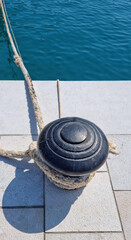 Fototapeta premium A thick nautical rope is tied around a black cast iron mooring bollard on a stone pier in Baska on the island of Krk with blue sea water in the background