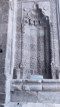 Vertical static shot of a man walking across and out of the frame in front of an ornate ancient stone portal. Showcases scale and historical atmosphere
