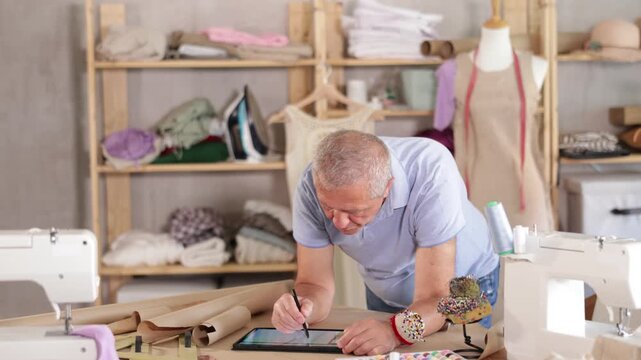 Elderly male tailor draws pattern using tablet in sewing workshop