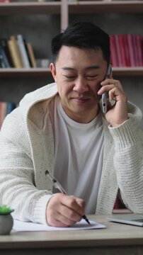 A man is recording a video of himself on a smartphone while talking on the phone and writing notes at his desk