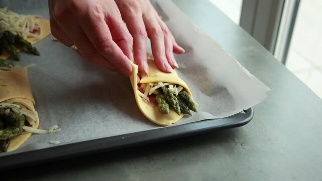 Close-up of wrapping raw puff pastry squares around prosciutto and fresh asparagus bundles with cheese on a baking tray
