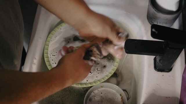 Man washes dishes in a kitchen sink with a sponge and liquid soap under running water