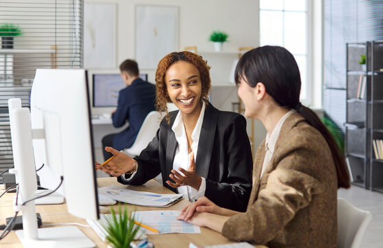 Businesswomen colleagues meeting teamwork office analytics discussion. Smiling colleagues talk at a computer, review reports, share insights, and align steps. Clear teamwork and planning idea.