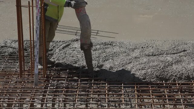 Construction worker in safety vest using a concrete pump to pour wet cement mixture over a steel reinforcement mesh, creating a solid building foundation on a construction site