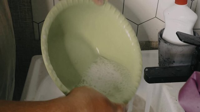 Man washes dishes in a kitchen sink with a sponge and liquid soap under running water.