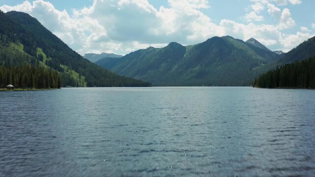 Drone flying straight over calm Rakhmanovskoye Lake surrounded by mountains and pine forest in Altai, East Kazakhstan. Wide alpine panorama with clear water and untouched wilderness scenery.