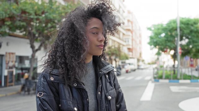 Teenage woman with curly hair touches cheek and smiles on a street, wearing jacket and sweater; playful youthful confidence.