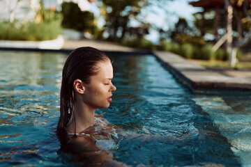 Fototapeta premium Young woman with wet hair relaxes in outdoor swimming pool during sunny day surrounded by trees and clear water lifestyle concept