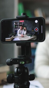 A man is recording a video of himself holding a small wooden block while sitting at a desk with a laptop and books in the background