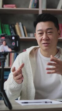 A man is actively gesturing while recording himself on a smartphone mounted on a tripod, with a laptop open on his desk