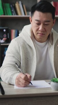 A man in a cozy cardigan is actively filming himself on a smartphone while taking notes at his desk, surrounded by books and a laptop