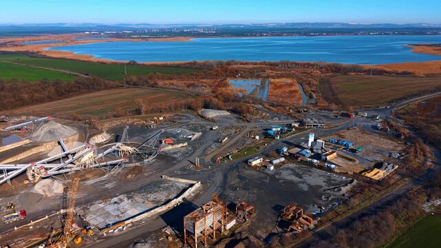 Wide aerial view of aggregate plant near blue lake. Industrial landscape featuring a crushing and screening plant with water reservoir, fields, and mountains in background.