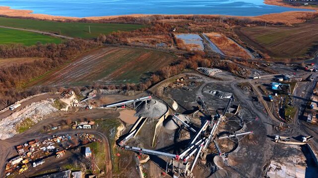 High angle aerial view of gravel quarry near lake. Industrial mining facility with stone piles and conveyor belts located next to agricultural fields and a large lake.